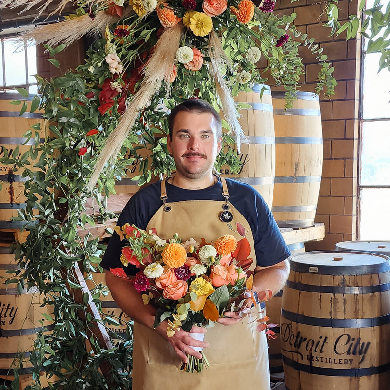 Founder Michael Bak-Ferrer standing in front of a floral arch, holding a bouquet of bright, colorful mixed flowers.